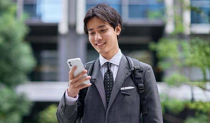 屋外でスマートフォンを操作しながら笑顔で見つめるスーツ姿の男性