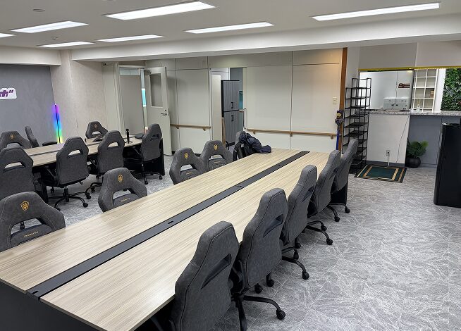 天井にLED照明が並ぶ広々とした研修室。木目調の天板にグレーのゲーミングチェアを合わせたデスクが3列並び、各デスク中央に電源コンセントが設置されている。左奥のグレーの壁には紫と青のLEDストリップライト、正面奥の壁には複数の白い扉と黒いロッカー、2つのシューズラックが見える。