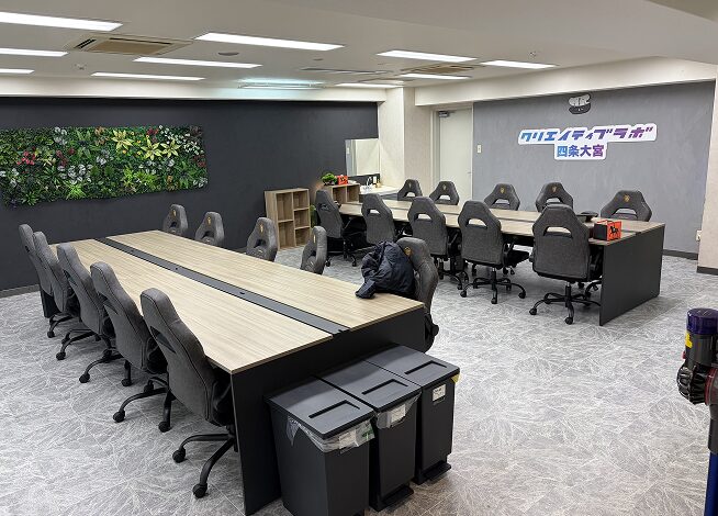 手前から奥へ続く木目調のデスクとグレーのゲーミングチェア、右手前に置かれたグレーのゴミ箱。奥の黒い壁には大きなウォールグリーンが飾られ、その右隣のグレーの壁には「クリエイティブラボ 四条大宮」と書かれた看板が設置されている。